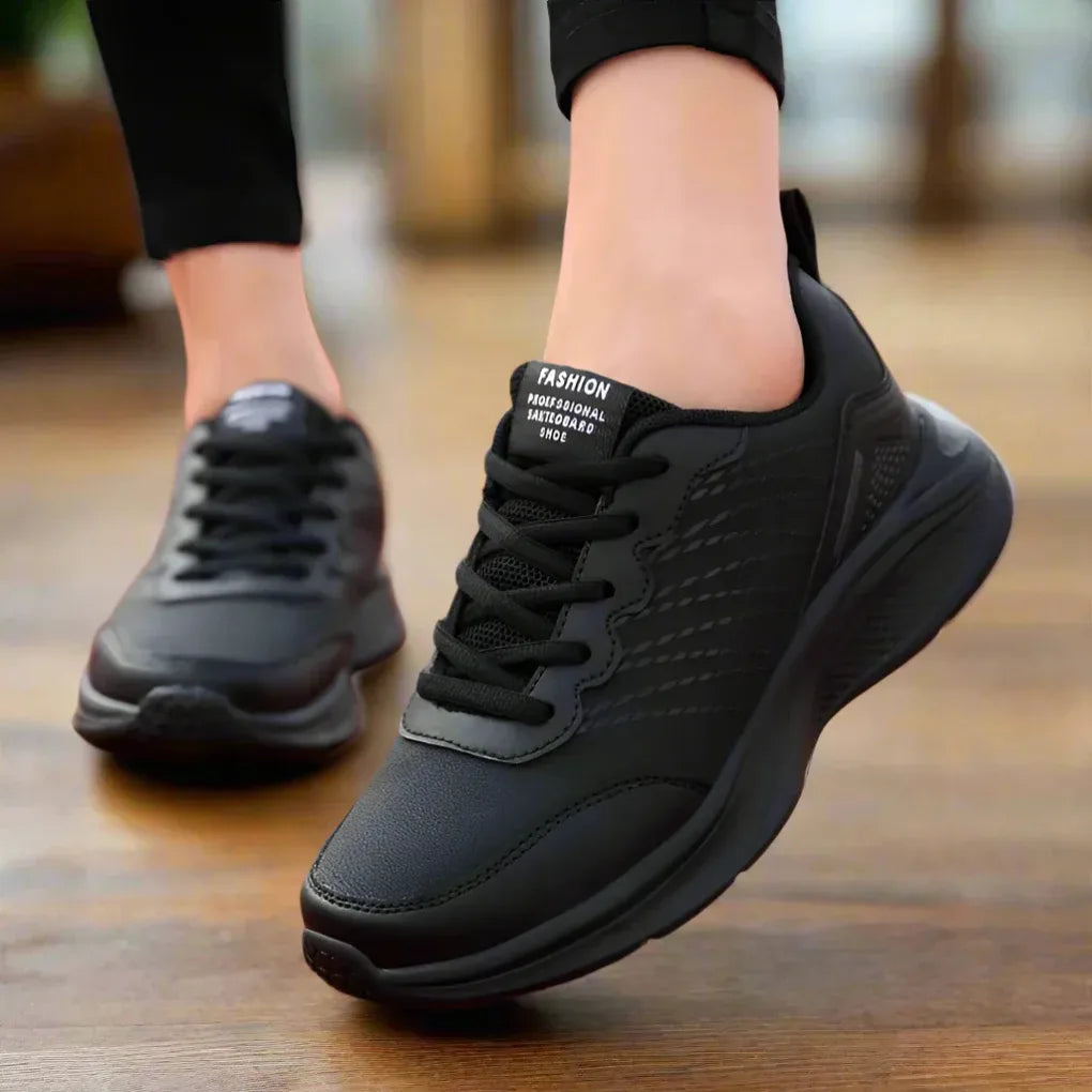 Close-up side view of a black lace-up trainer with textured panelling and a thick black midsole, worn while walking indoors.