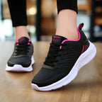 Black and pink lace-up trainer with a chunky white midsole and pink trim on the heel and tongue, shown mid-stride on a wooden floor.