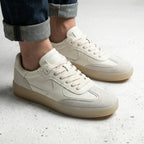 Side view of a man wearing beige leather trainers with suede panels and rolled-up jeans on a concrete floor.