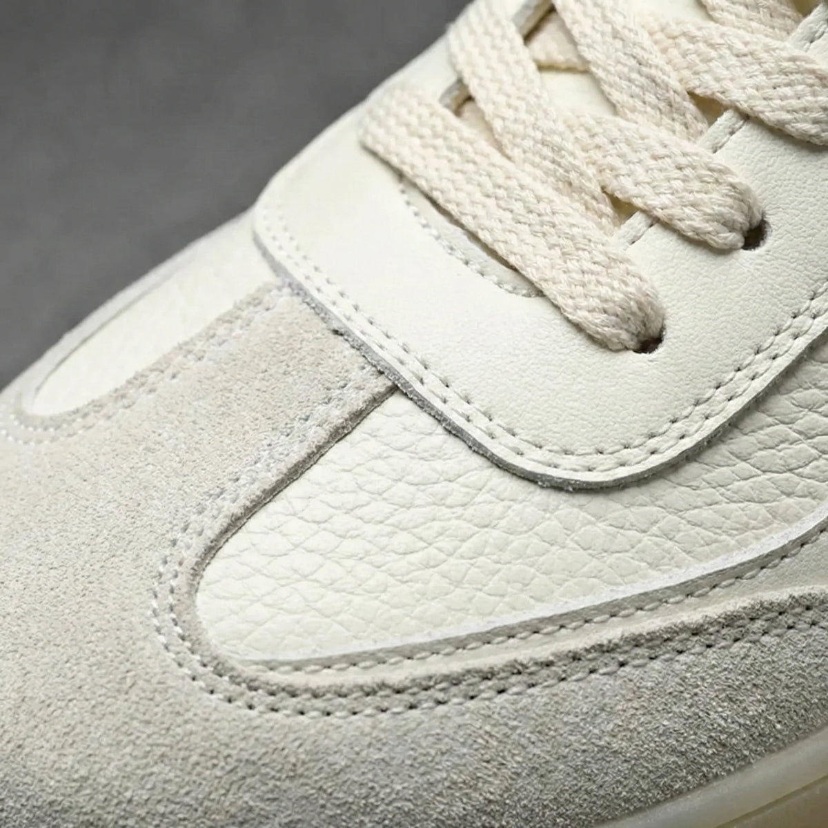 Close-up of the beige trainer's toe box showing stitched suede and leather panels with cream laces.