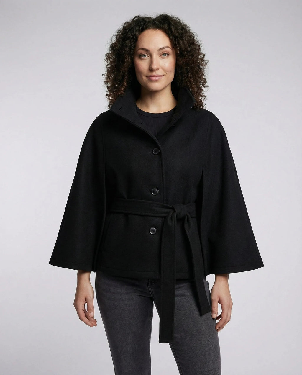 Front view of a belted black wool-blend cape jacket with stand collar, worn over a black top and grey skinny jeans