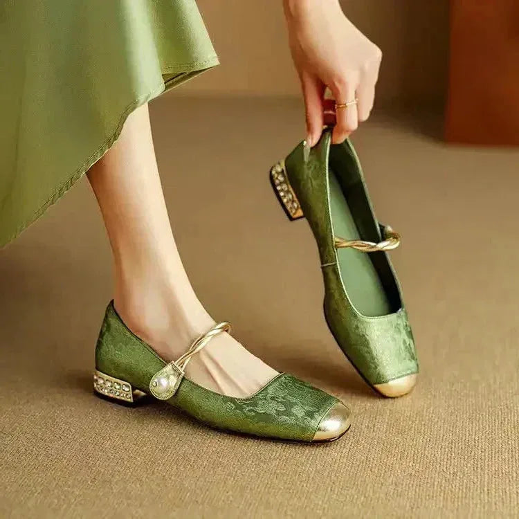 Close-up of a hand holding a green brocade Mary Jane flat with gold toe cap and braided pearl strap
