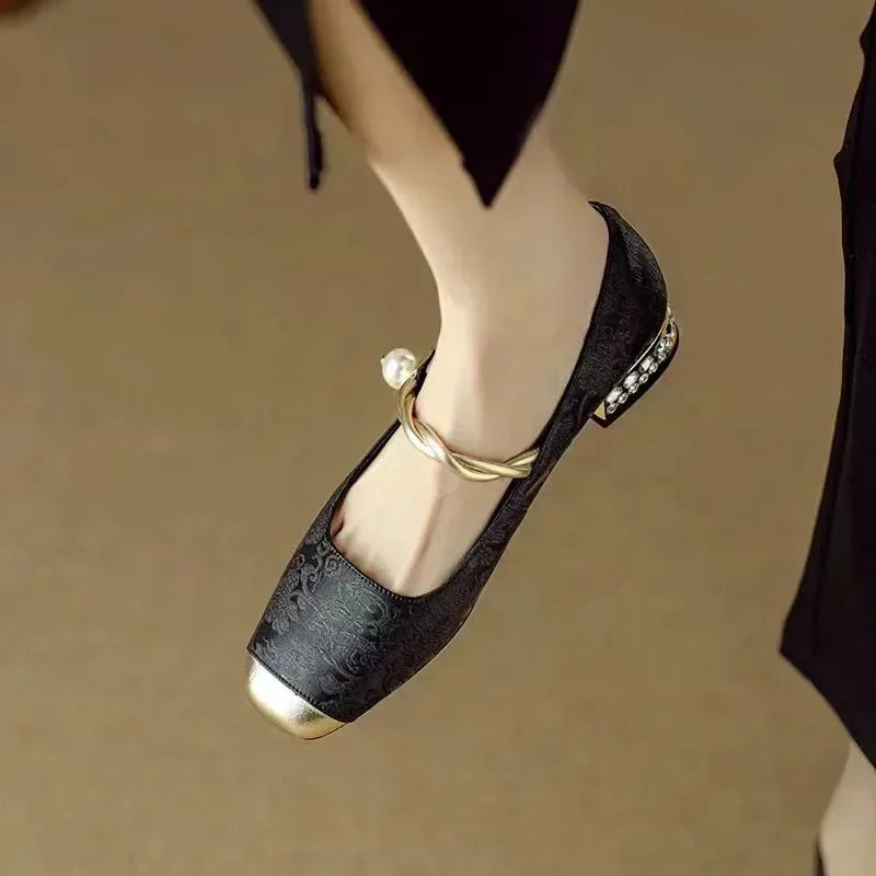 Close-up of a black brocade Mary Jane flat with gold toe cap worn on the ankle, twisted pearl strap visible