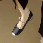 Close-up of a black brocade Mary Jane flat with gold toe cap worn on the ankle, twisted pearl strap visible
