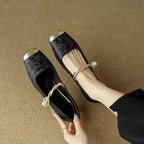 Overhead shot of a pair of black brocade flats held in hand, showing the braided pearl ankle strap