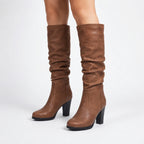 Side view of brown vegan leather knee-high boots with slouchy ruched shaft and block heel, worn on bare legs.
