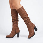 Walking side view of the brown knee-high boots showing the gathered calf, pointed toe and stacked block heel.