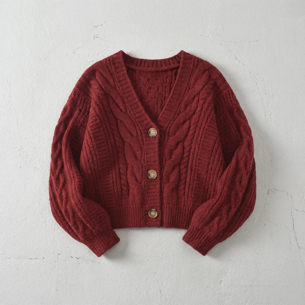 Deep red cable-knit cropped cardigan with wooden buttons and balloon sleeves, laid flat on a light stone backdrop