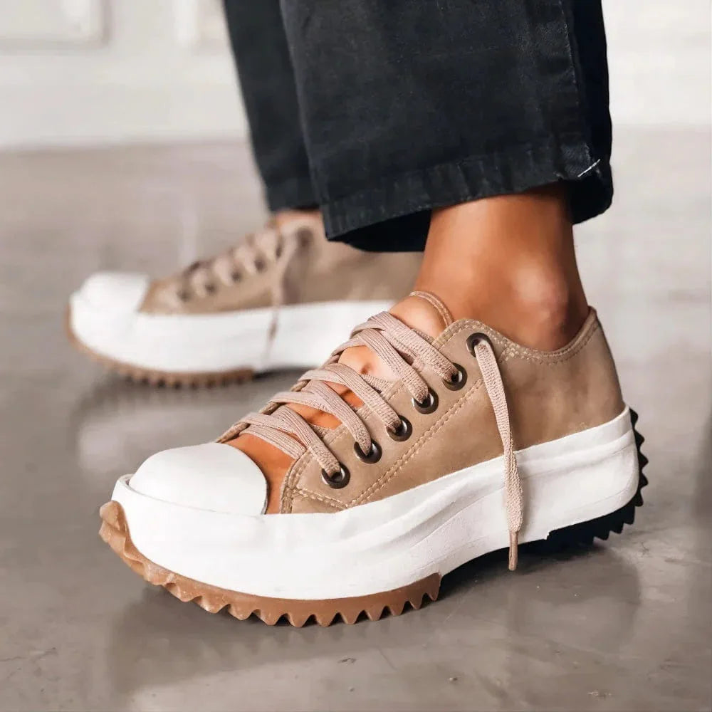 Side view of beige suede platform trainer with white toe cap and chunky tread sole, worn with dark trousers.