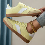 Pale yellow retro trainer held in hand showing white side stripe, suede toe cap and gum rubber sole in profile view