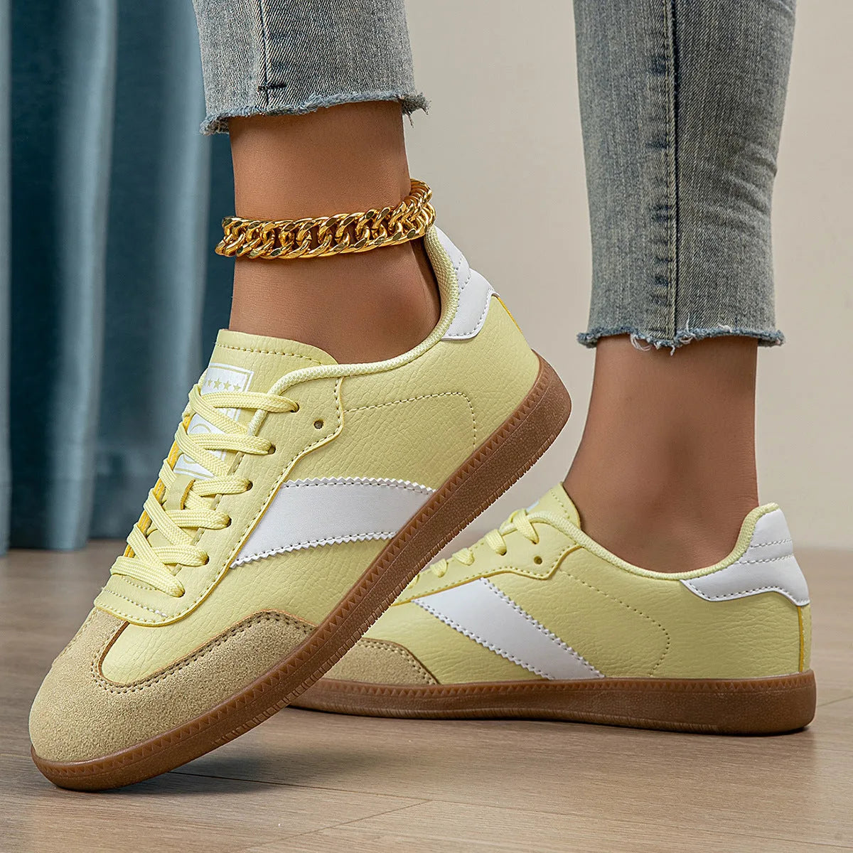 Woman wearing pale yellow lace-up trainers with white panel detail and gold chain anklet, standing on wooden floor