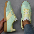 Pair of mint green trainers with pale yellow stripe and laces, one worn on foot, one held showing side profile and sole