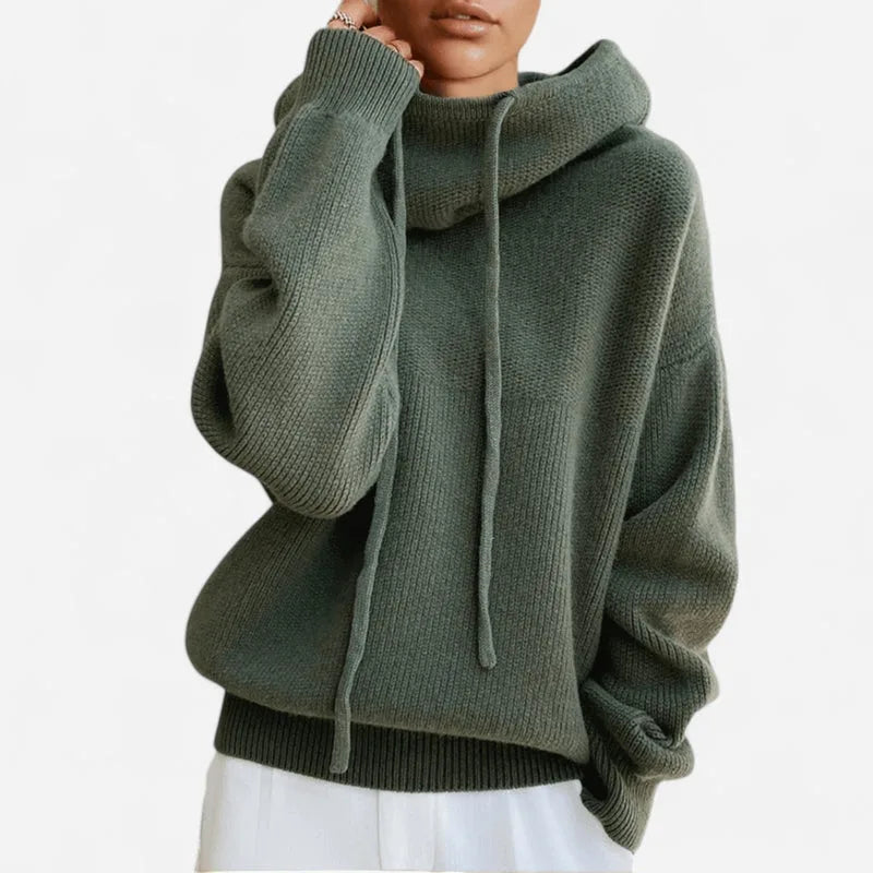 Close-up front view of a green chunky knit cowl neck jumper with drawstring hood worn with white trousers