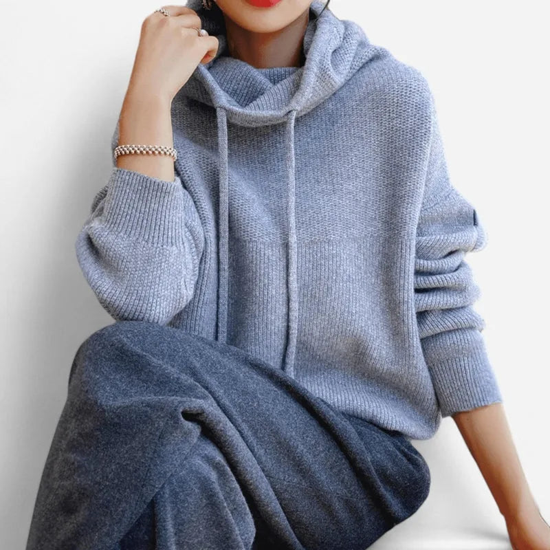 Woman seated wearing a light blue cowl neck knitted hoodie with drawstring detail and pearl bracelet