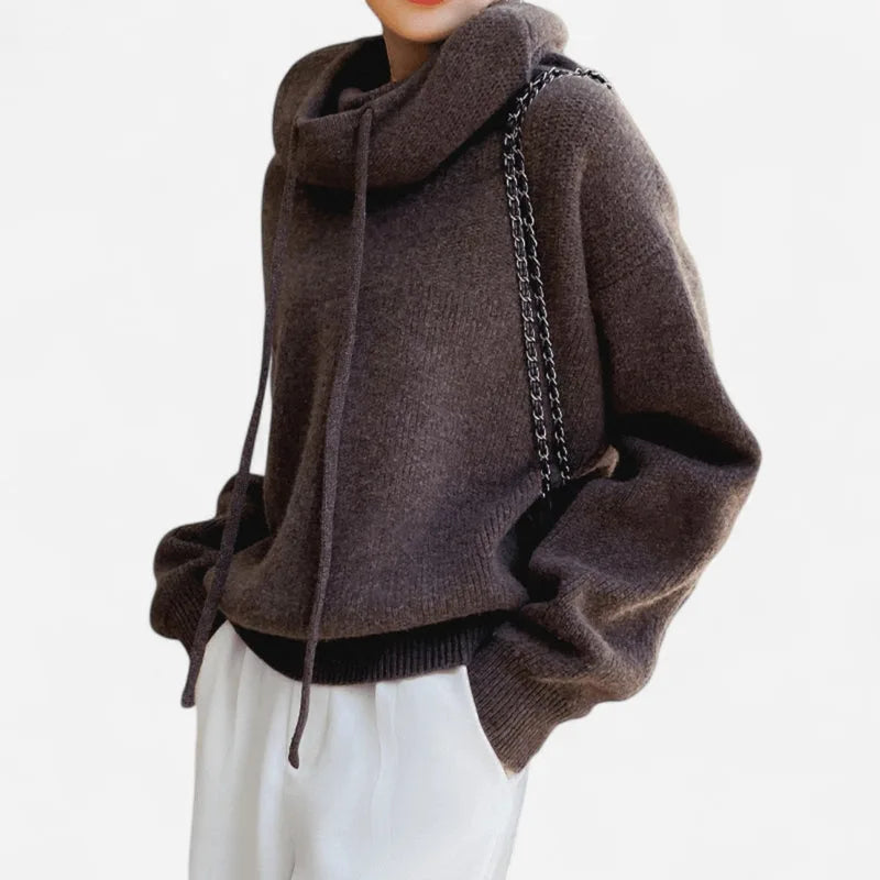 Side view of a brown chunky knit cowl neck hoodie paired with white trousers and chain bag strap