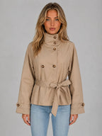 Front view of model wearing a beige cropped trench jacket with tortoiseshell buttons and a tied waist belt over blue jeans