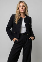 Woman in a collarless black tweed cropped jacket with gold buttons and flap pockets over black trousers