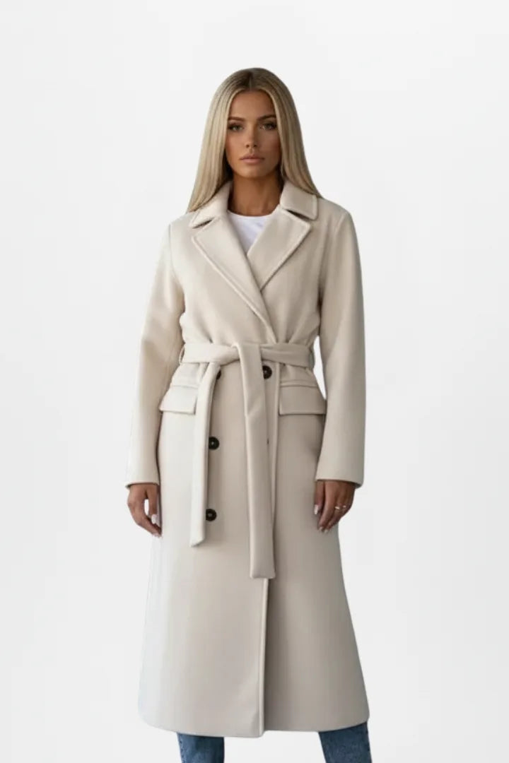 Front view of a woman wearing a beige double-breasted longline coat with tie belt and notch collar over jeans