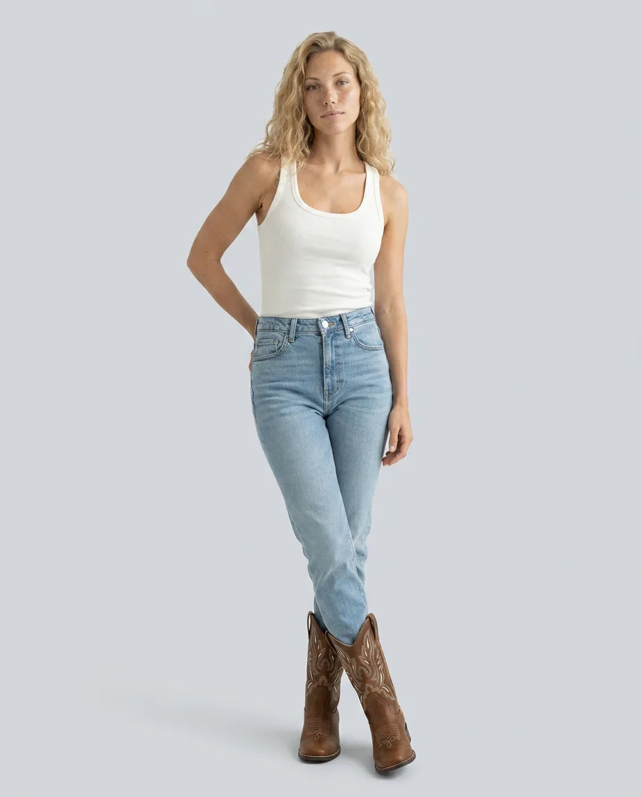 Woman in white vest top and jeans styled with brown western boots, standing against a plain background