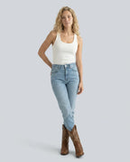 Woman in white vest top and jeans styled with brown western boots, standing against a plain background