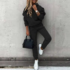 Woman in an oversized black hooded tracksuit with drawstring hood, elastic waist joggers and white trainers, holding a black handbag