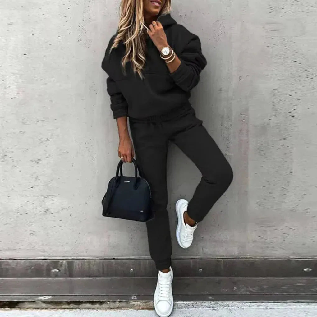 Woman in an oversized black hooded tracksuit with drawstring hood, elastic waist joggers and white trainers, holding a black handbag