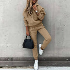 Camel-toned fleece hoodie and jogger tracksuit worn with white trainers, shown against a concrete wall
