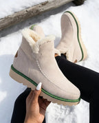 Beige suede platform ankle boot with cream faux fur trim, held by hand against a snowy log backdrop