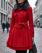 Close-up of a woman in a red double-breasted skater coat with a plush faux fur collar and bow-tie waist belt, standing on a cobbled street