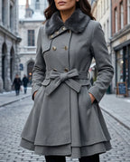 Grey wool-blend belted coat with a dark faux fur collar and bronze buttons, shown with hands in the front pockets