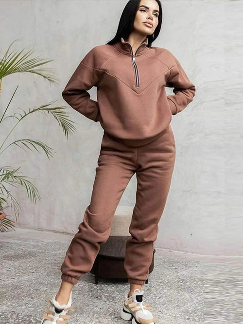 Woman wearing a brown half-zip fleece tracksuit with tapered joggers and white trainers, styled with a palm plant backdrop.