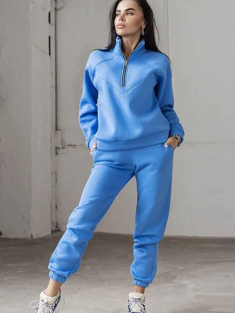 Front view of a bright blue half-zip sweatshirt and matching jogger set worn with hands in pockets against a grey wall.