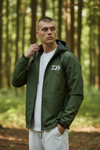 Man in a forest wearing an open khaki green hooded jacket over a white t-shirt, holding the collar.