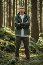 Man standing arms crossed wearing the khaki green hooded jacket zipped up, paired with dark green trousers outdoors.