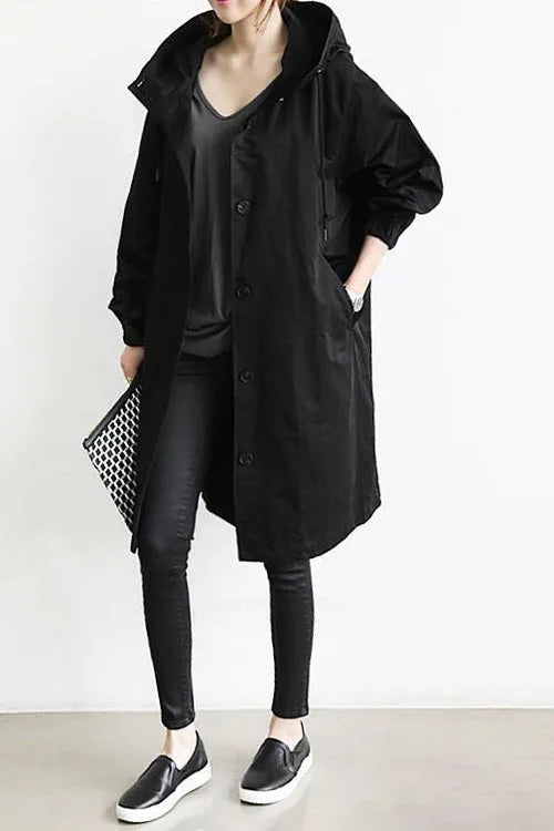 Black hooded longline coat worn with skinny trousers and slip-on trainers, styled with a patterned clutch bag