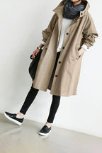 Beige hooded longline coat worn open over a white jumper, paired with black trousers and trainers