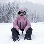 Woman sits in deep snow wearing a dusty pink hooded ski jacket, goggles and white gloves, pine forest behind