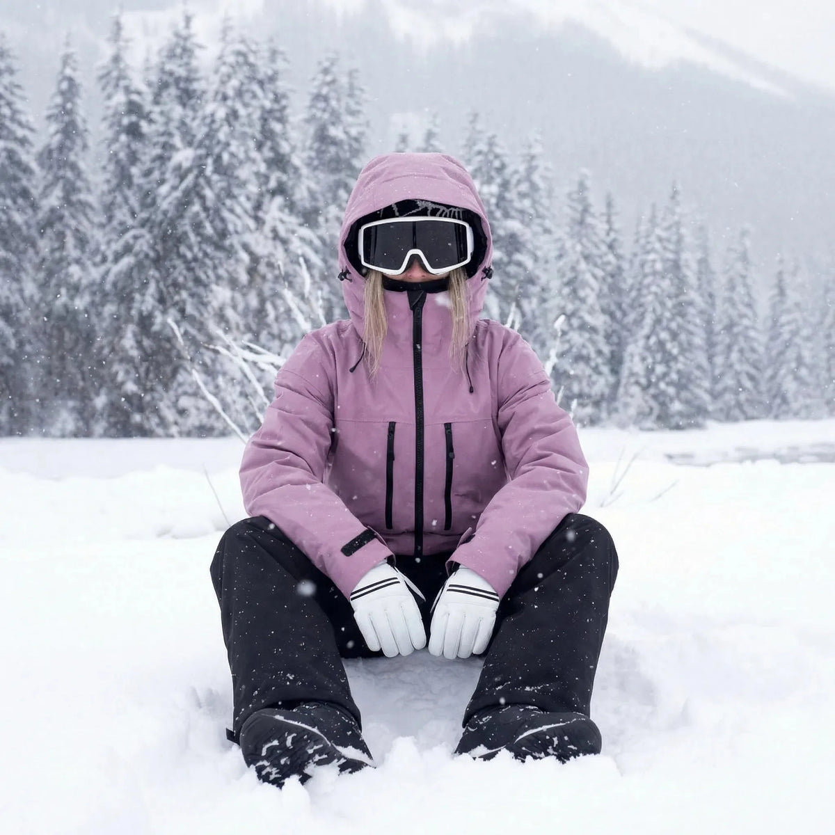 Woman sits in deep snow wearing a dusty pink hooded ski jacket, goggles and white gloves, pine forest behind