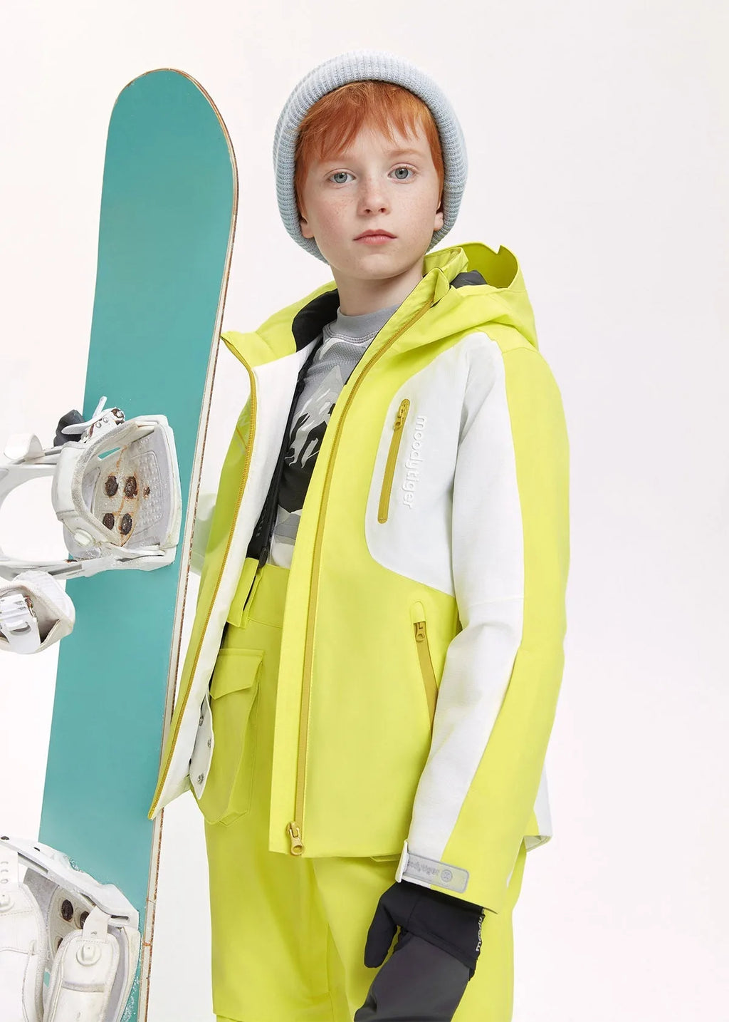 Boy wearing a lime yellow and white ski jacket with grey beanie, standing beside a teal snowboard