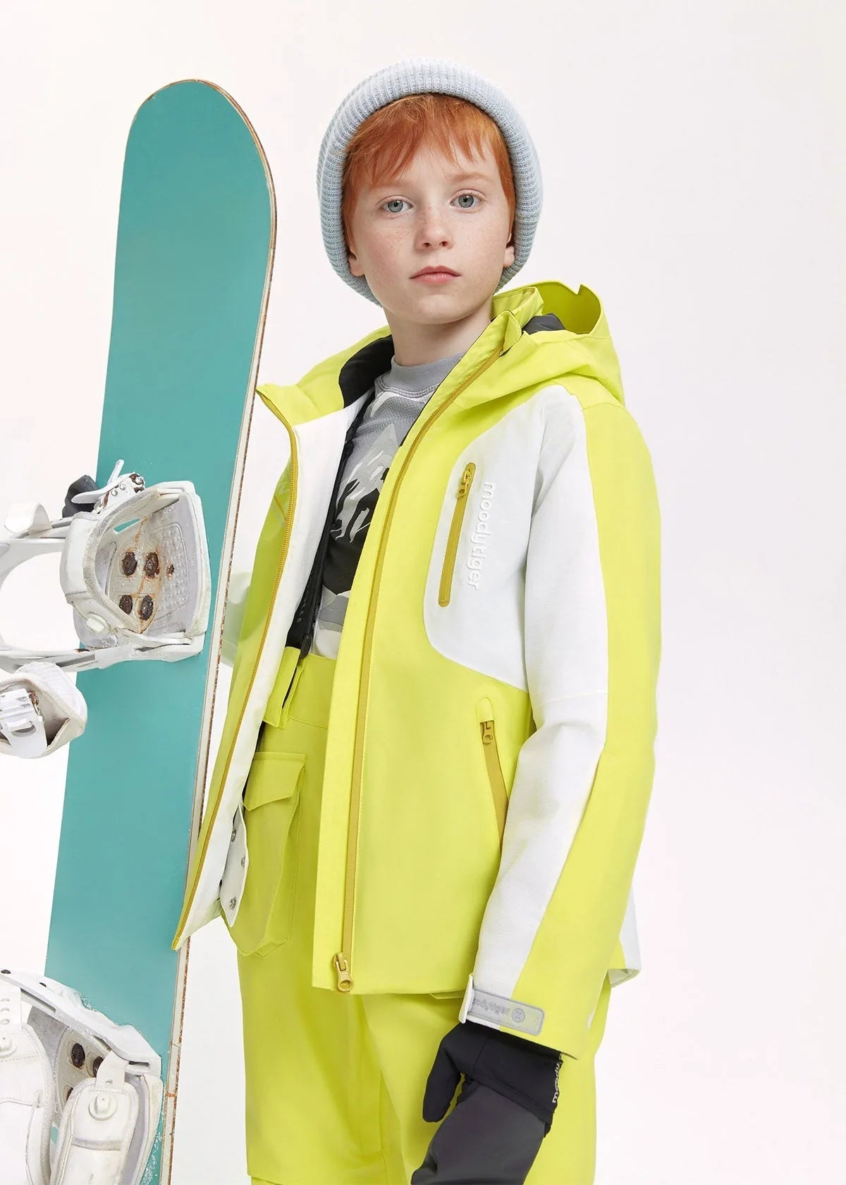 Boy wearing a lime yellow and white ski jacket with grey beanie, standing beside a teal snowboard