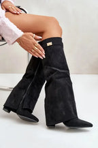 Model seated wearing black suede pointed-toe flap boots with gold buckle accents
