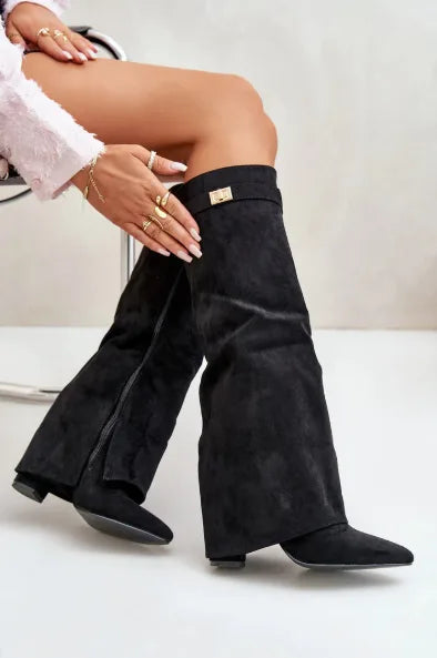 Model seated wearing black suede pointed-toe flap boots with gold buckle accents
