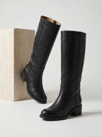 Pair of black leather knee-high boots with block heel, one leaning against a stone block, side view