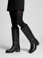 Woman wearing black leather knee-high boots with sheer black tights and a black skirt, walking pose