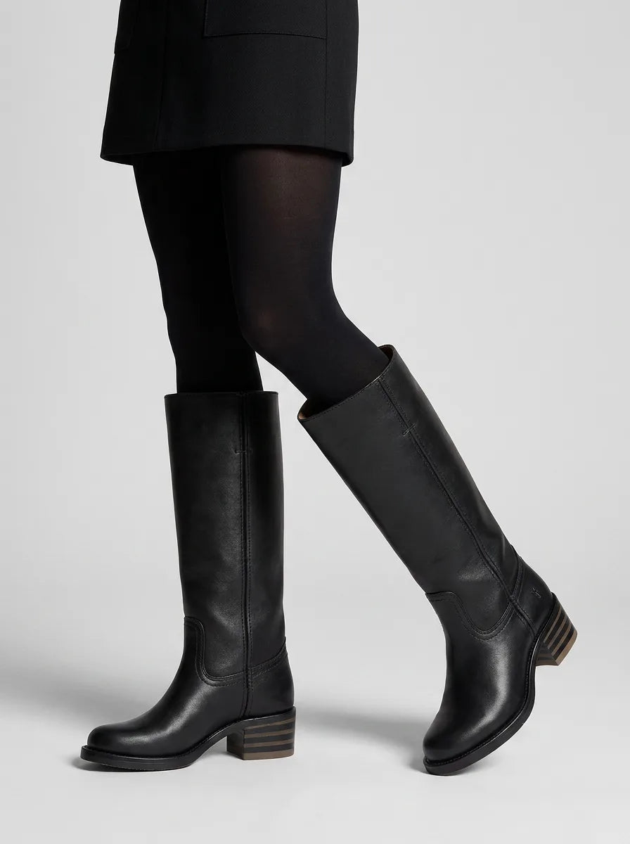 Woman wearing black leather knee-high boots with sheer black tights and a black skirt, walking pose