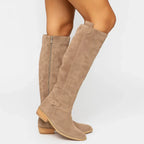 Side view of beige suede knee-high boots with a side zip, buckle strap and low stacked block heel