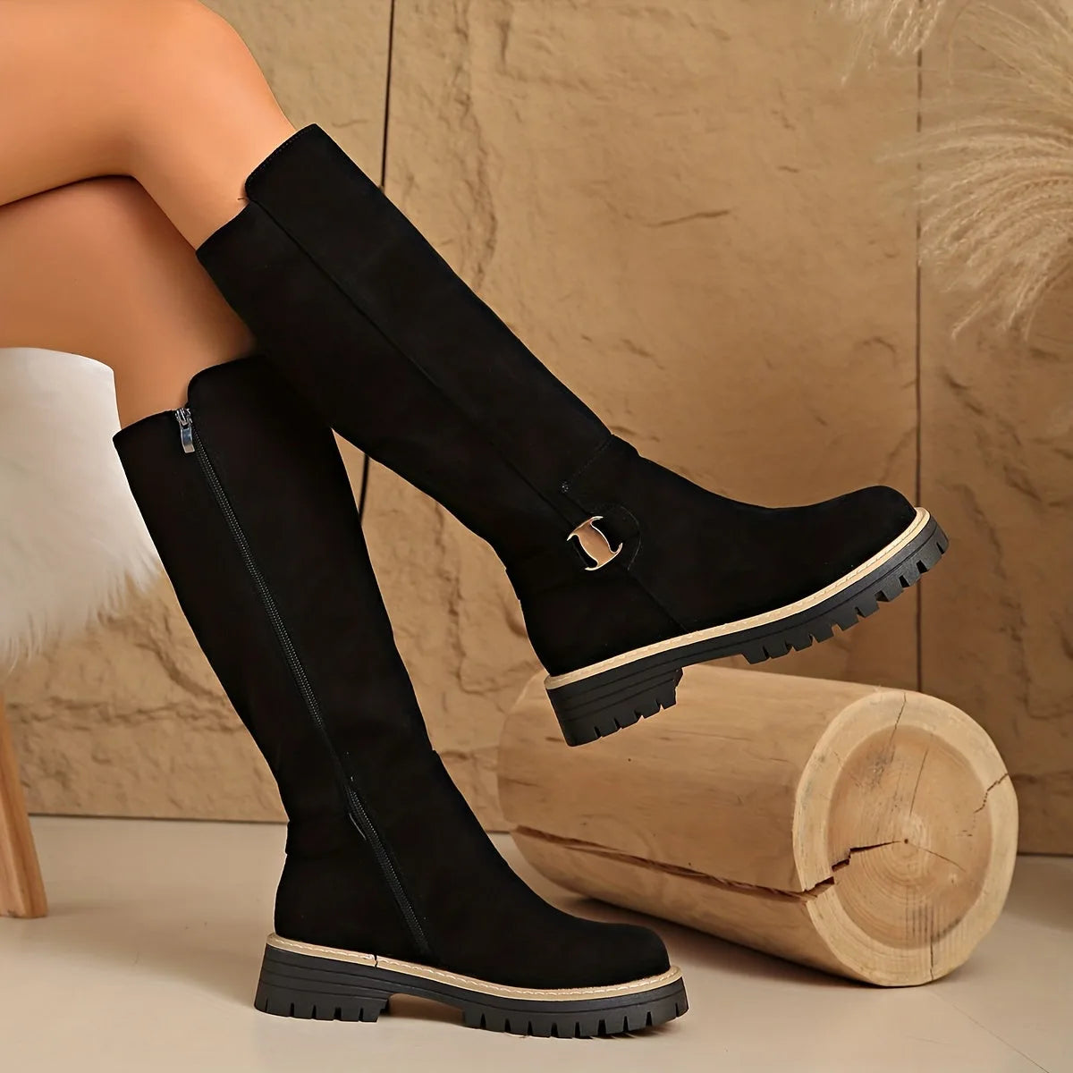 Side view of black suede knee-high boots with chunky lug sole and ankle buckle, worn with legs crossed