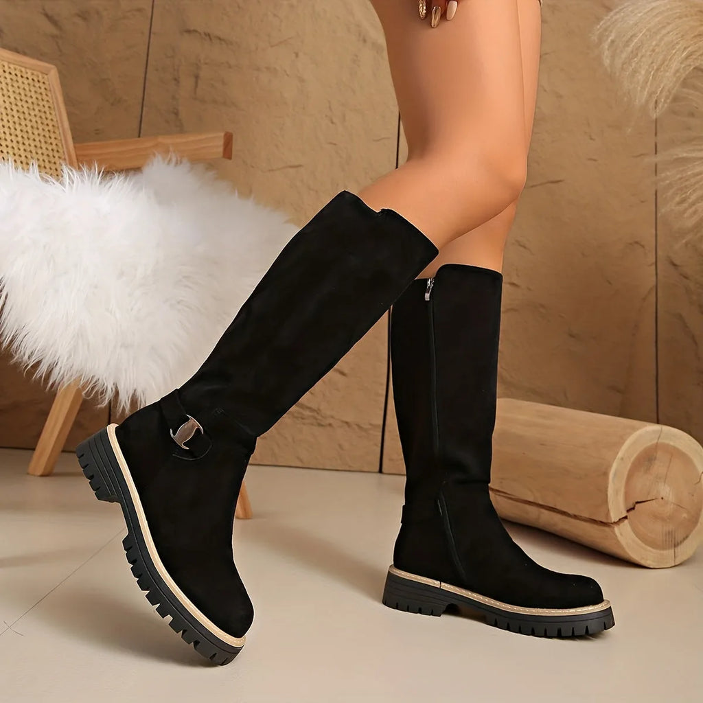 Pair of black suede knee-high boots with ridged platform sole, one boot resting on a wooden log