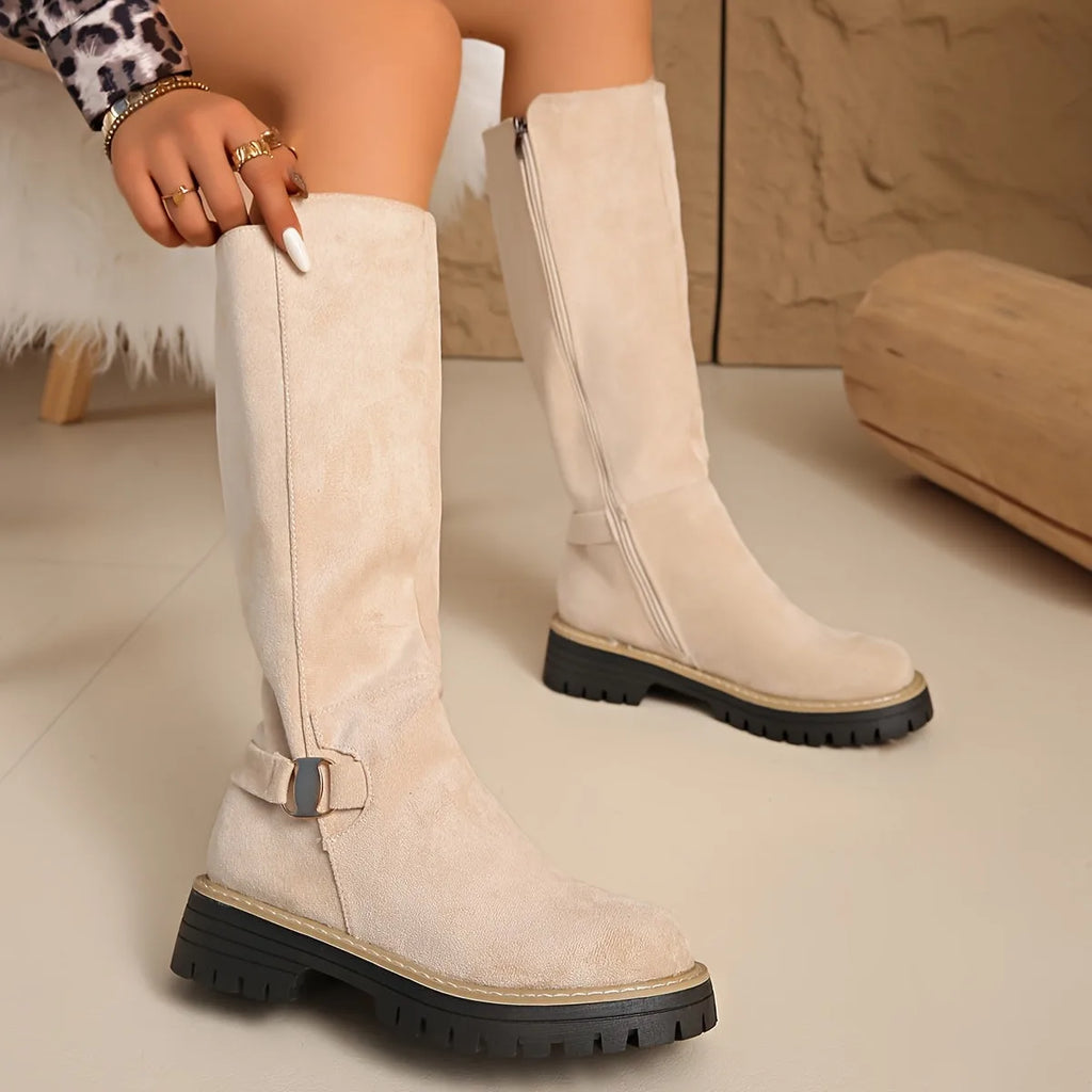 Close-up of beige suede knee-high boot with side zip and metal buckle detail, hand adjusting the shaft