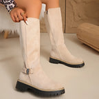 Close-up of beige suede knee-high boot with side zip and metal buckle detail, hand adjusting the shaft